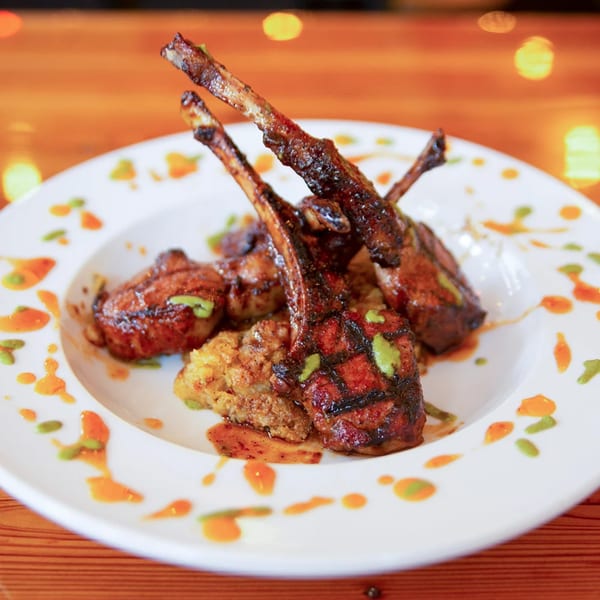 Grilled lamb chops with a side of rice, drizzled with orange and green sauces on a white plate.