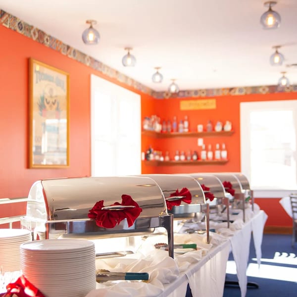 A buffet line with several chafing dishes, plates, and serving utensils in a bright orange room.