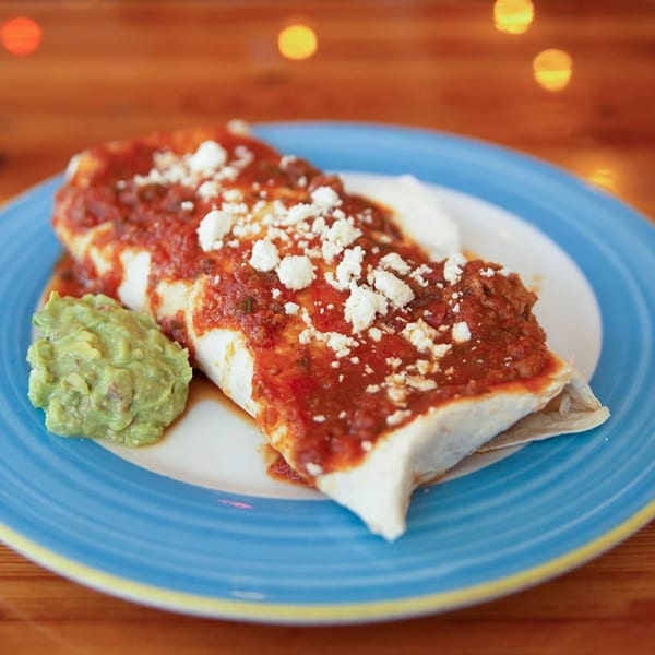 A Mexican burrito covered in red sauce and crumbled cheese, served with guacamole and sour cream.