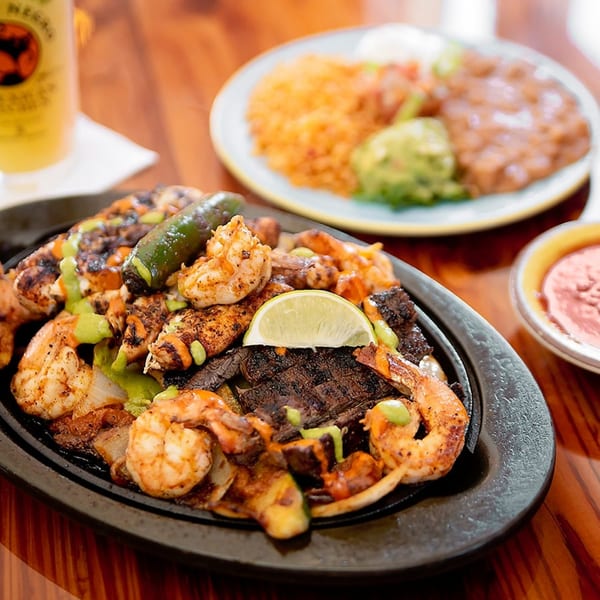 Sizzling fajitas with shrimp, steak, chicken, peppers, onions, and lime, with rice and beans in the background.