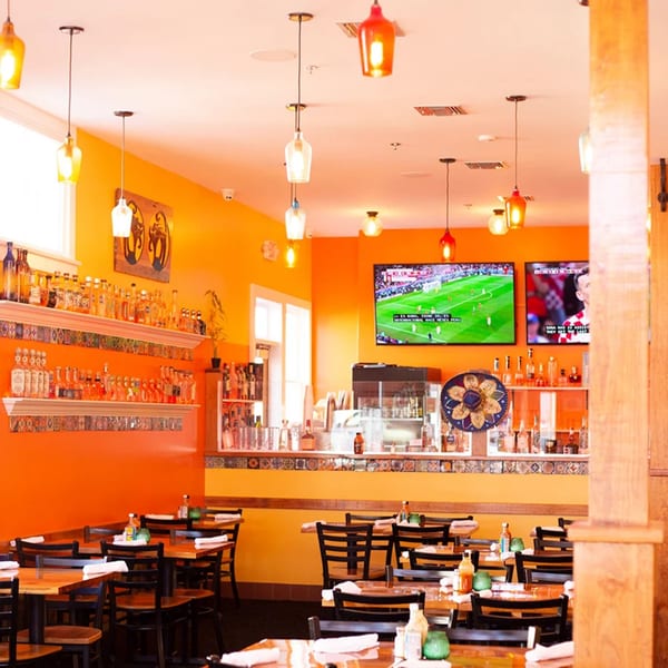Vibrant orange interior of El Gato Negro Mexican restaurant with tables, bar, and TVs showing soccer.