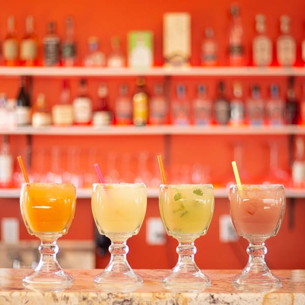 Four colorful margaritas with straws and salted rims on a bar with a red background.
