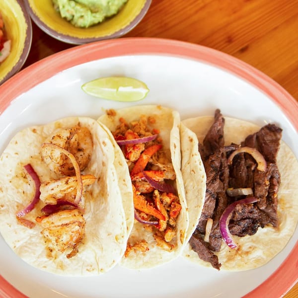 Three El Gato Negro tacos, including shrimp, chicken, and steak, with a lime wedge and guacamole.