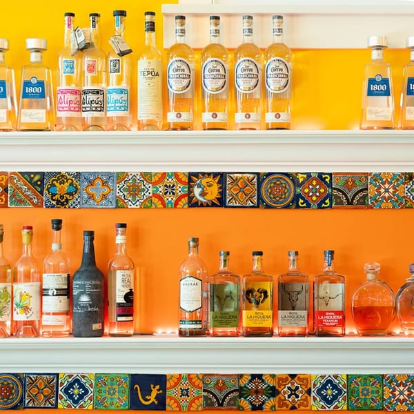 Two shelves of various tequila and mezcal bottles against a vibrant orange and yellow wall with decorative tiles.