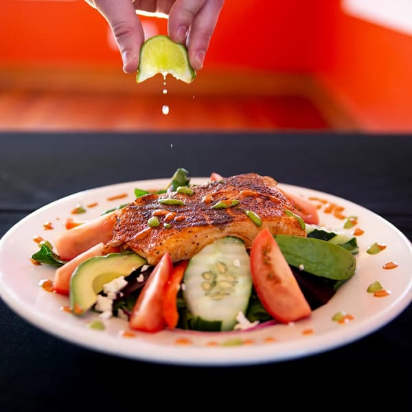A hand squeezes lime juice over a Mexican grilled salmon salad with avocado, tomatoes, and cucumbers.