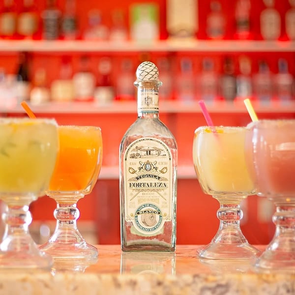 A bottle of Fortaleza tequila surrounded by four colorful margaritas on a bar.