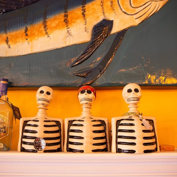 Three decorative skeleton figures and a tequila bottle on a shelf, with a large fish carving above.