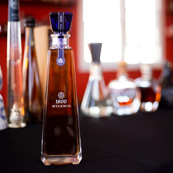 A bottle of 1800 Milenio tequila stands on a dark surface, with other bottles blurred in the background.